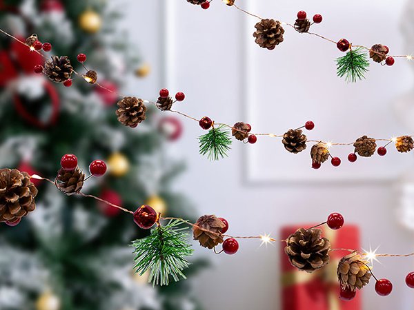 Christmas garland with pinecones, red berries, bells, and pine sprigs, illuminated by warm white LED lights, with a blurred Christmas tree in the background.