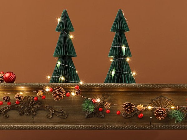Two green honeycomb Christmas trees with warm white LED lights, displayed on a mantelpiece with pinecone and berry garland.