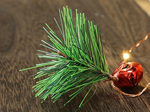 Close-up of a prelit Christmas garland section with pine needles, a red bell ornament, and warm white LED lights on a wooden surface.