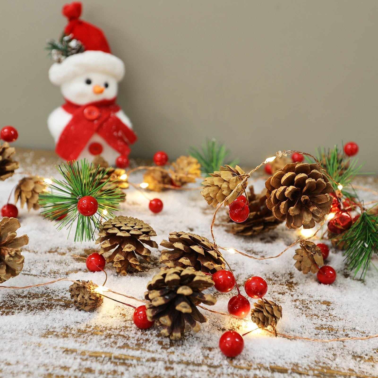 Christmas garland with pinecones, red berries, and LED lights scattered on a snowy wooden surface with a snowman in the background.