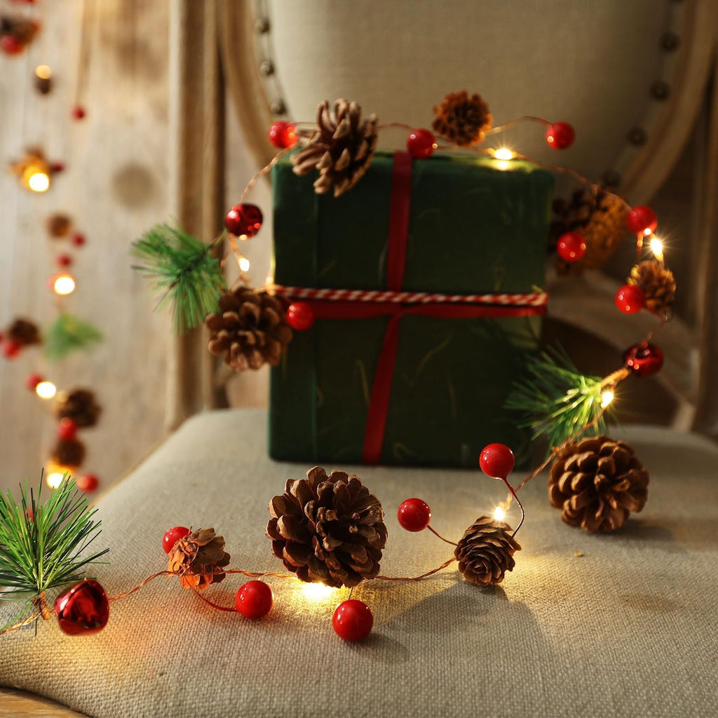 Christmas garland with pinecones, red berries, bells, and warm white LED lights draped around a gift.