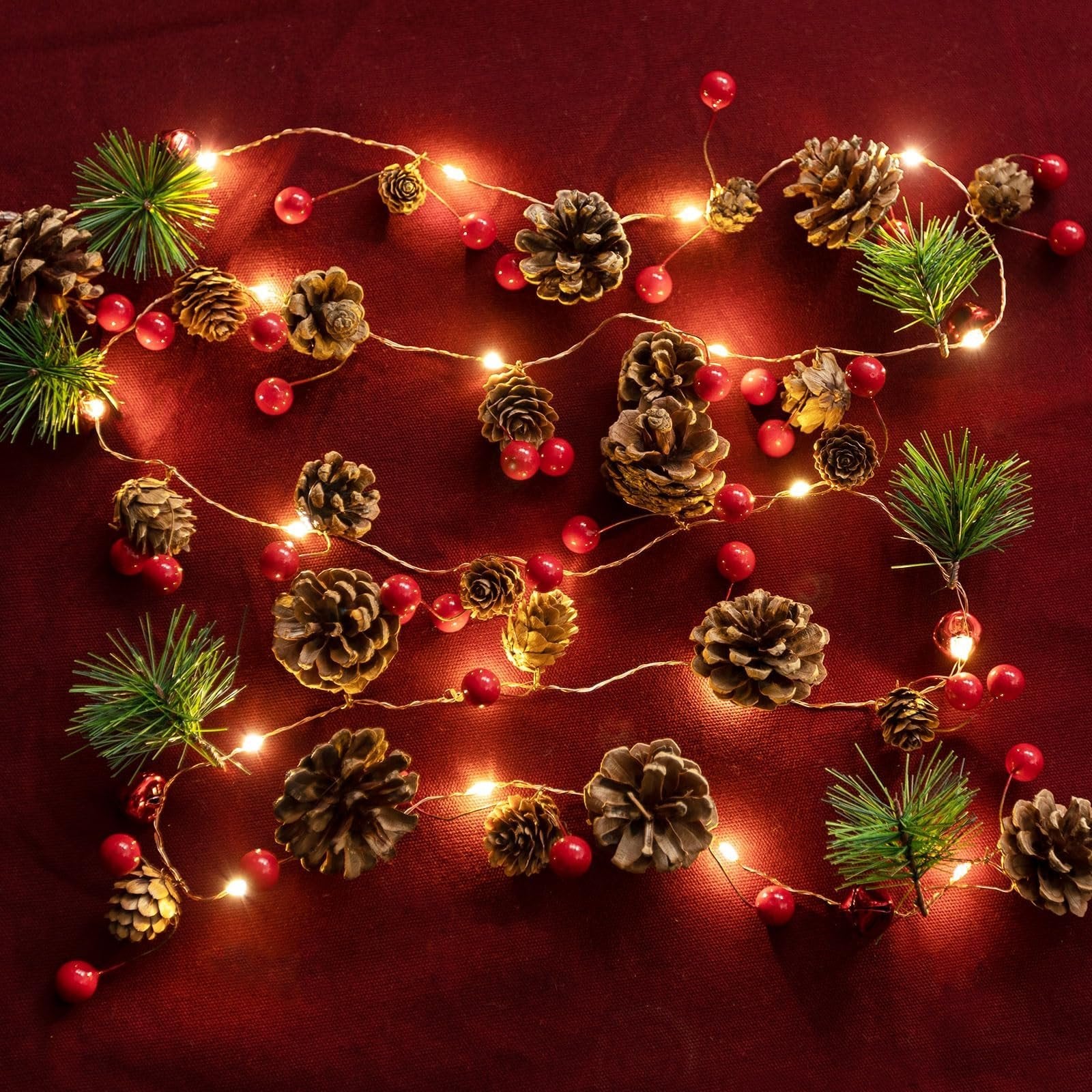 Christmas garland with pinecones, red berries, pine sprigs, and warm white LED lights on a red textured background.