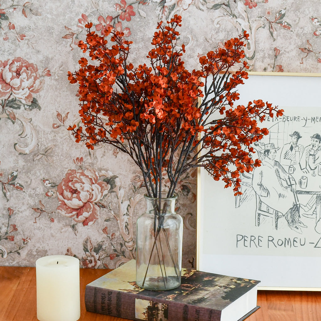Burnt orange artificial baby's breath flowers in a glass vase on a wooden table with a candle and a framed drawing.