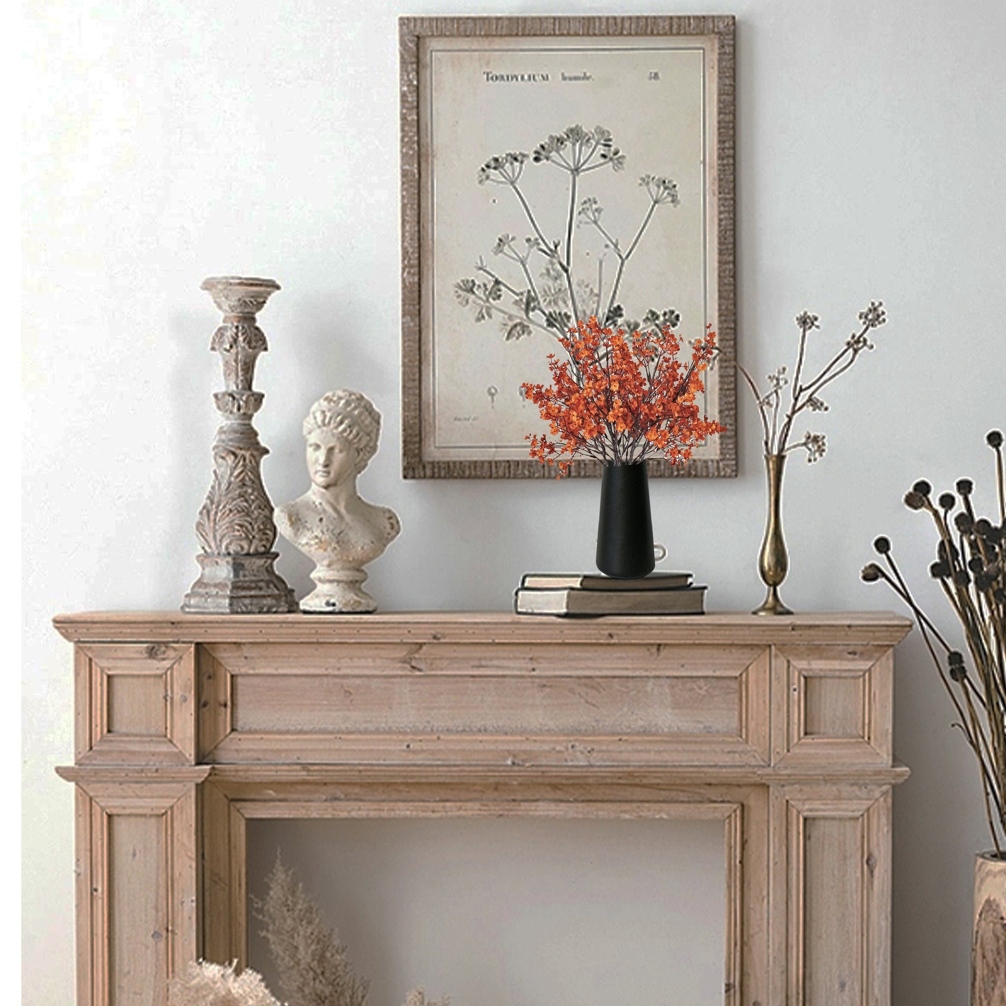 Burnt orange artificial baby's breath flowers in a black vase on a wooden mantelpiece with decorative items.