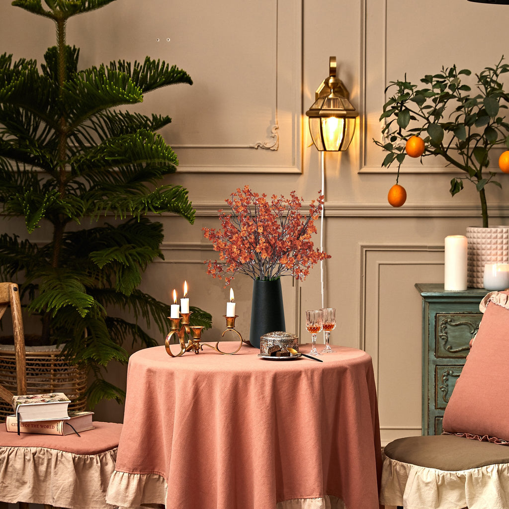 Burnt orange artificial baby's breath flowers in a dark vase on a pink tablecloth, with books and candles.