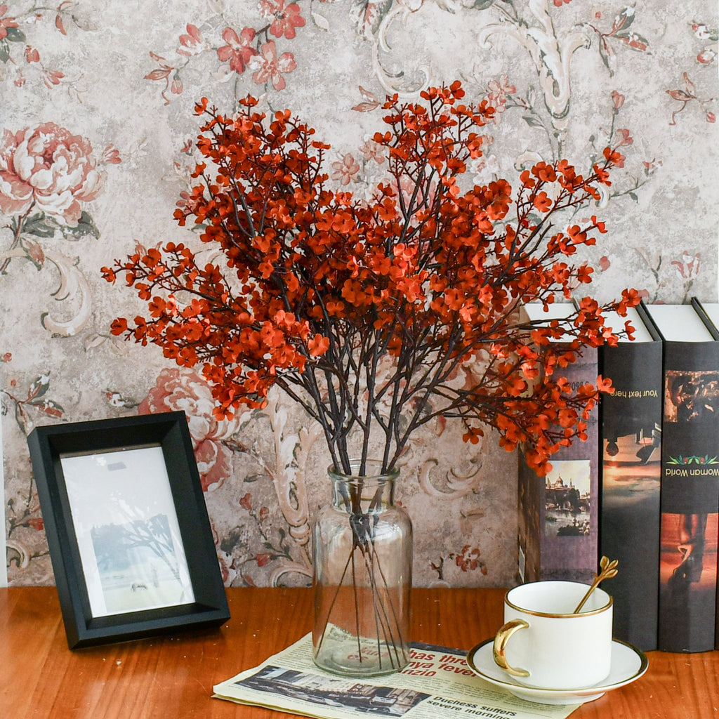 Burnt orange artificial baby's breath flowers in a glass vase on a wooden table with books and a teacup.