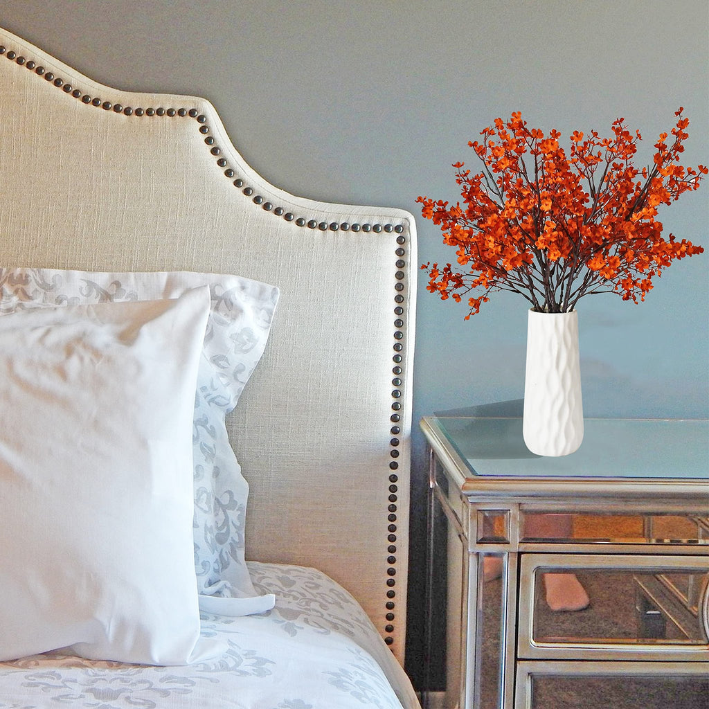 Burnt orange artificial baby's breath flowers in a white vase on a mirrored nightstand next to a tufted headboard.