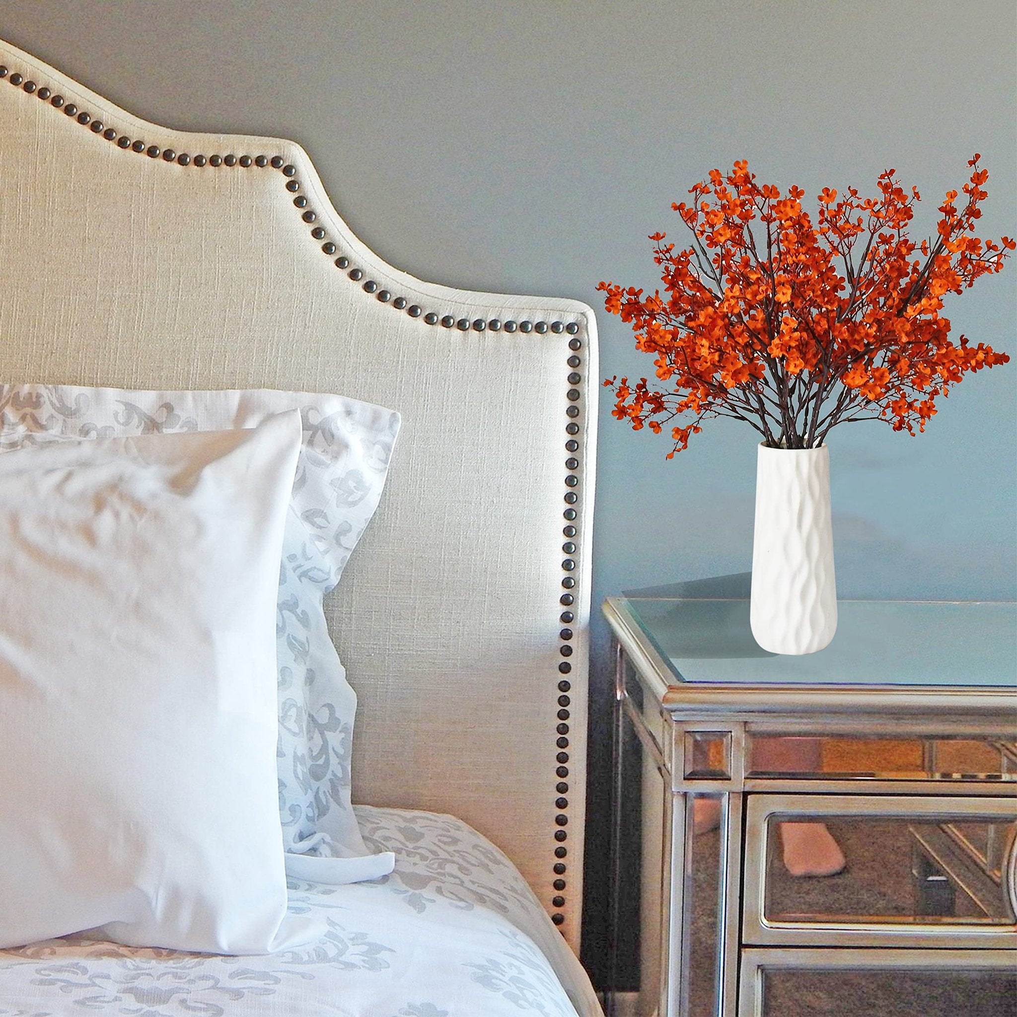 Burnt orange artificial baby's breath flowers in a white vase on a mirrored nightstand next to a tufted headboard.