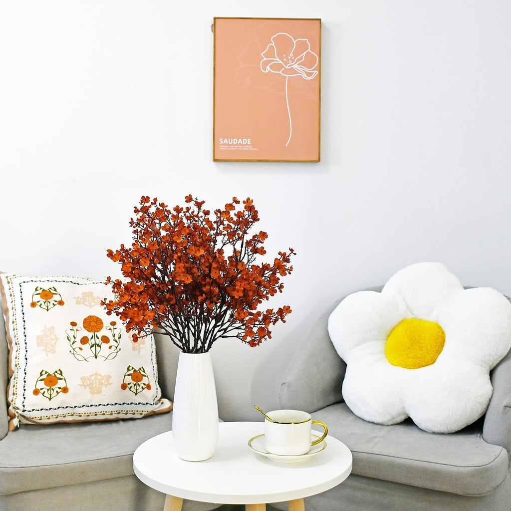 Burnt orange artificial baby's breath flowers in a white vase on a table, next to a couch with pillows and a teacup.