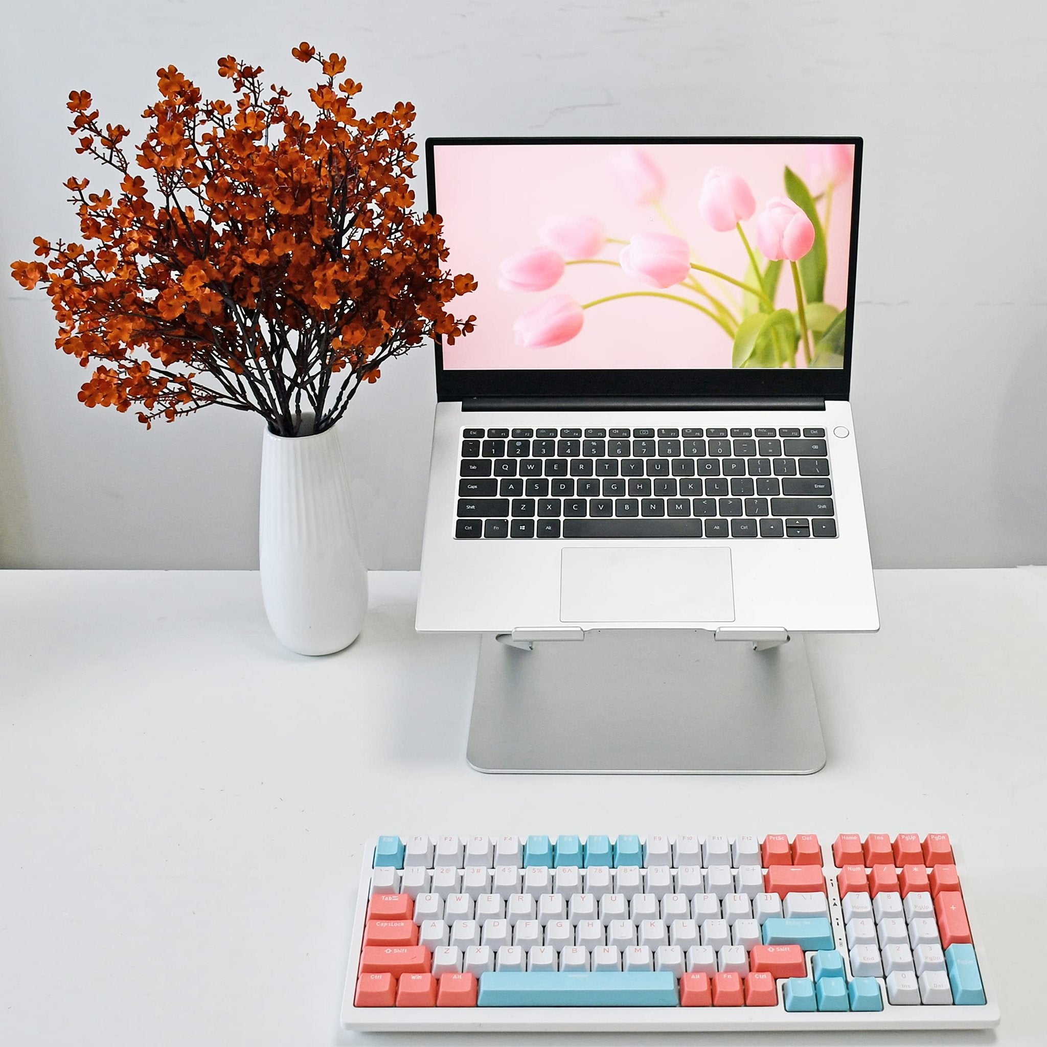 Burnt orange artificial baby's breath flowers in a white vase next to a laptop displaying pink tulips and a colorful keyboard.