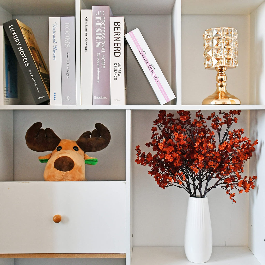 Burnt orange artificial baby's breath flowers in a white vase on a shelf next to books and a decorative lamp.