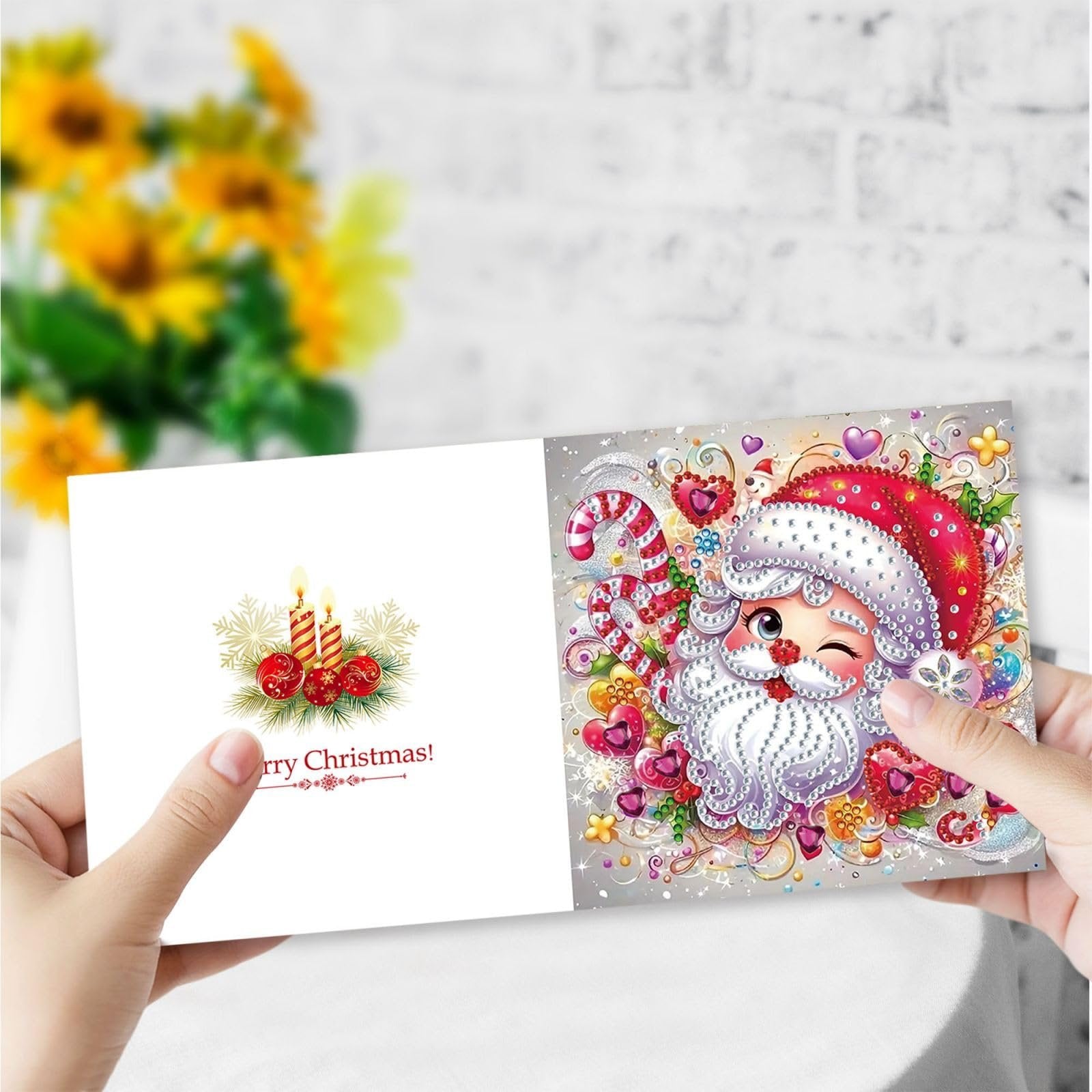 Hands holding open a Christmas greeting card with a diamond art Santa Claus design on the right and candles on the left.