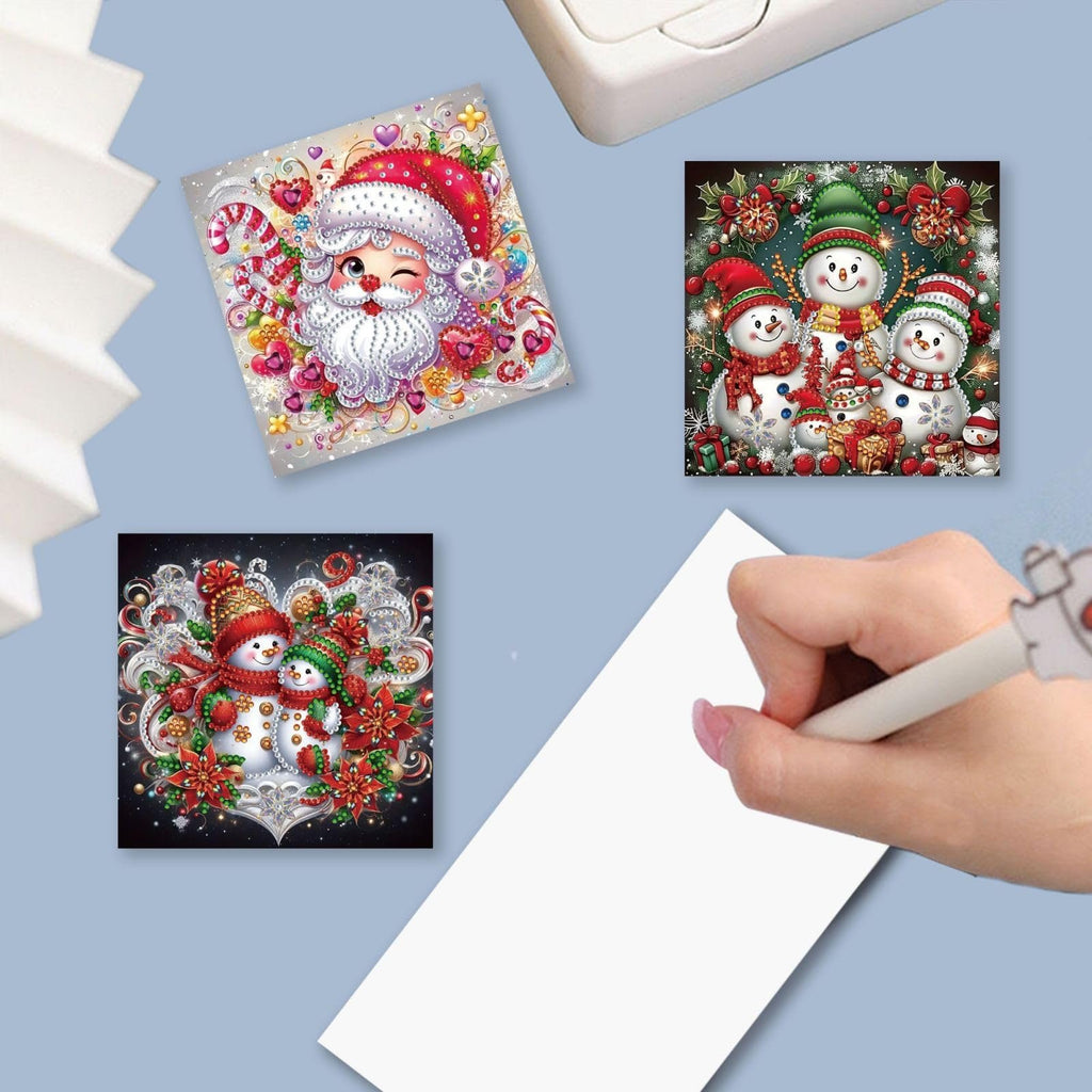 Diamond art Christmas cards featuring Santa and snowmen, with a hand writing on blank paper.
