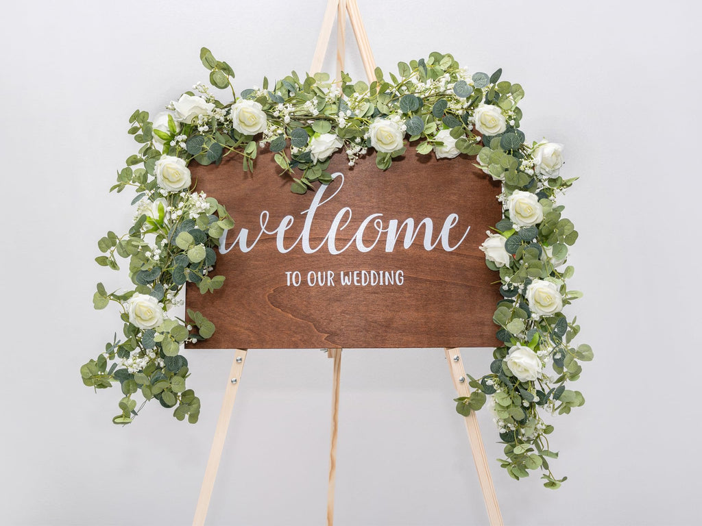 Wooden wedding welcome sign decorated with artificial eucalyptus garland and white roses, displayed on an easel.