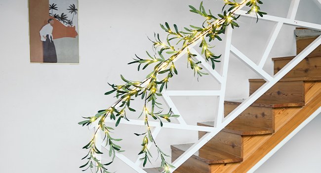 Artificial olive garland with warm white fairy lights draped along a white railing and wooden stairs
