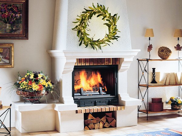 Lit artificial olive garland wreath above a fireplace with a fire burning, flanked by decor.