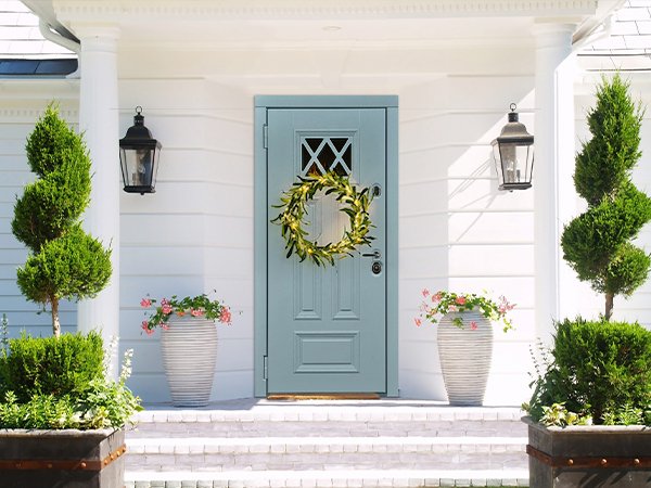 Lighted olive garland with warm white fairy lights on a blue front door, flanked by topiaries and planters.