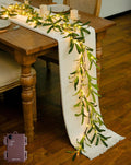 Artificial olive garland with warm white fairy lights draped on a table runner, with battery pack visible.