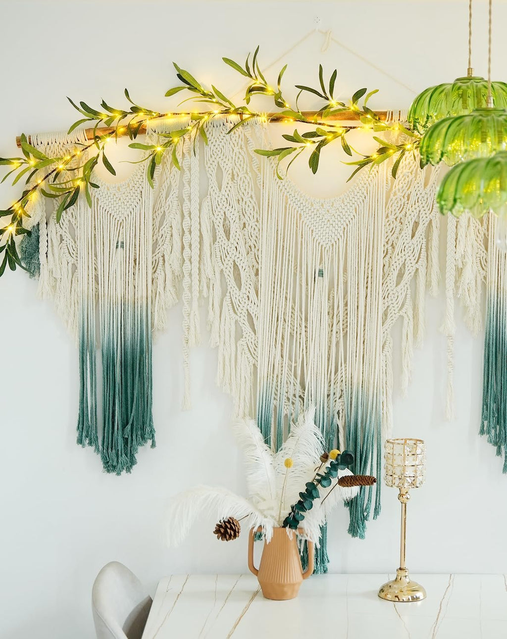 Macrame wall hanging with ombre green tassels and a lighted olive branch garland above.