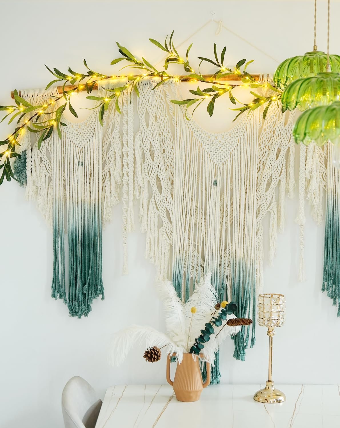 Macrame wall hanging with ombre green tassels and a lighted olive branch garland above.