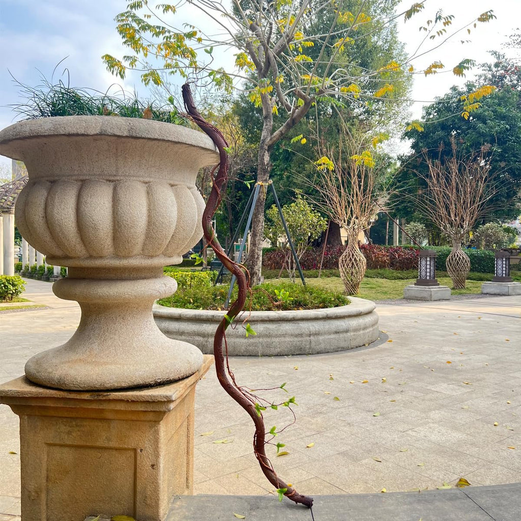 Artificial tree trunk vine with leaves draped over a stone planter in an outdoor garden setting.