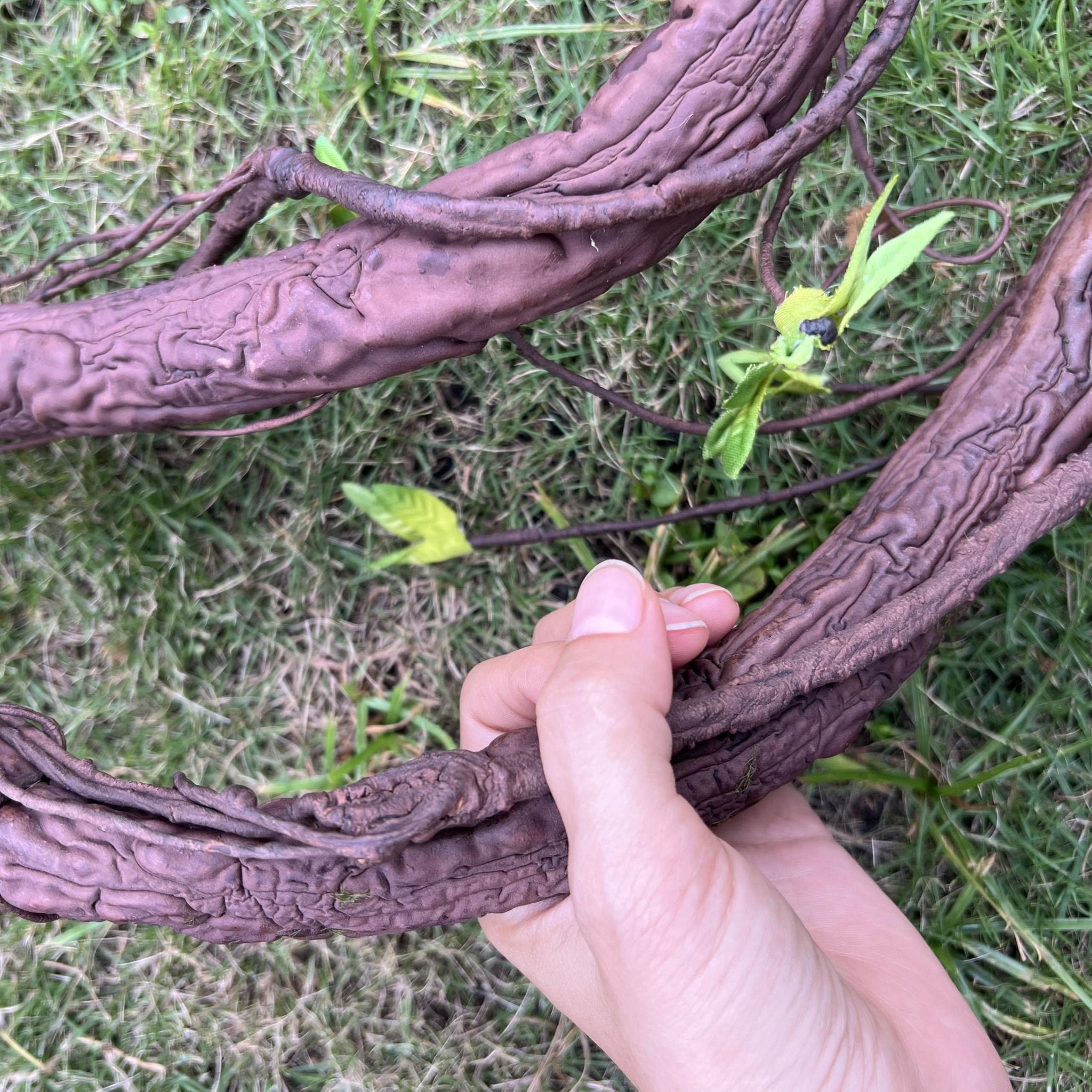 Hand holding a thick, bendable artificial tree trunk vine with textured bark and small green leaves on a grassy background.
