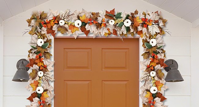 Autumn garland with maple leaves, white pumpkins, berries, and lights draped over a door.