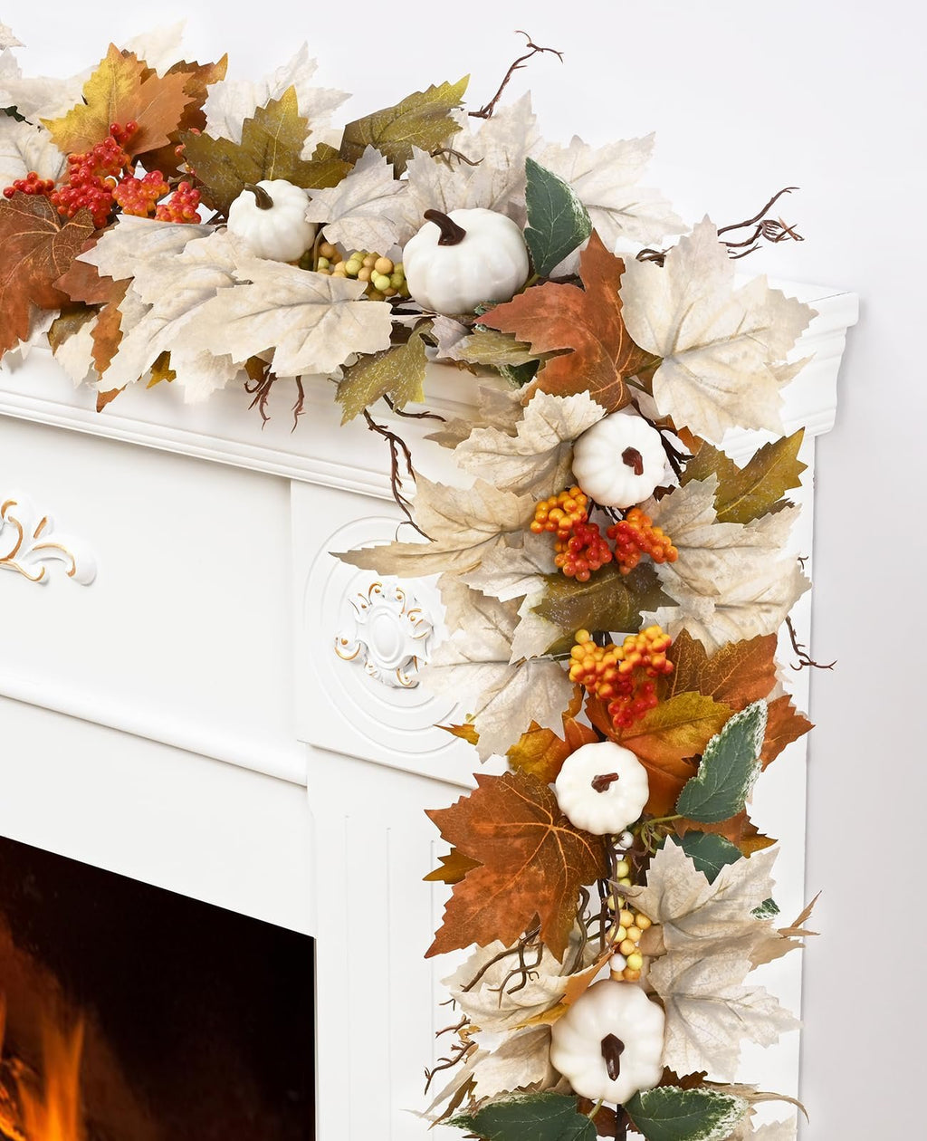 Autumn garland with maple leaves, white pumpkins, and berries draped over a white fireplace mantel with a fire visible below.