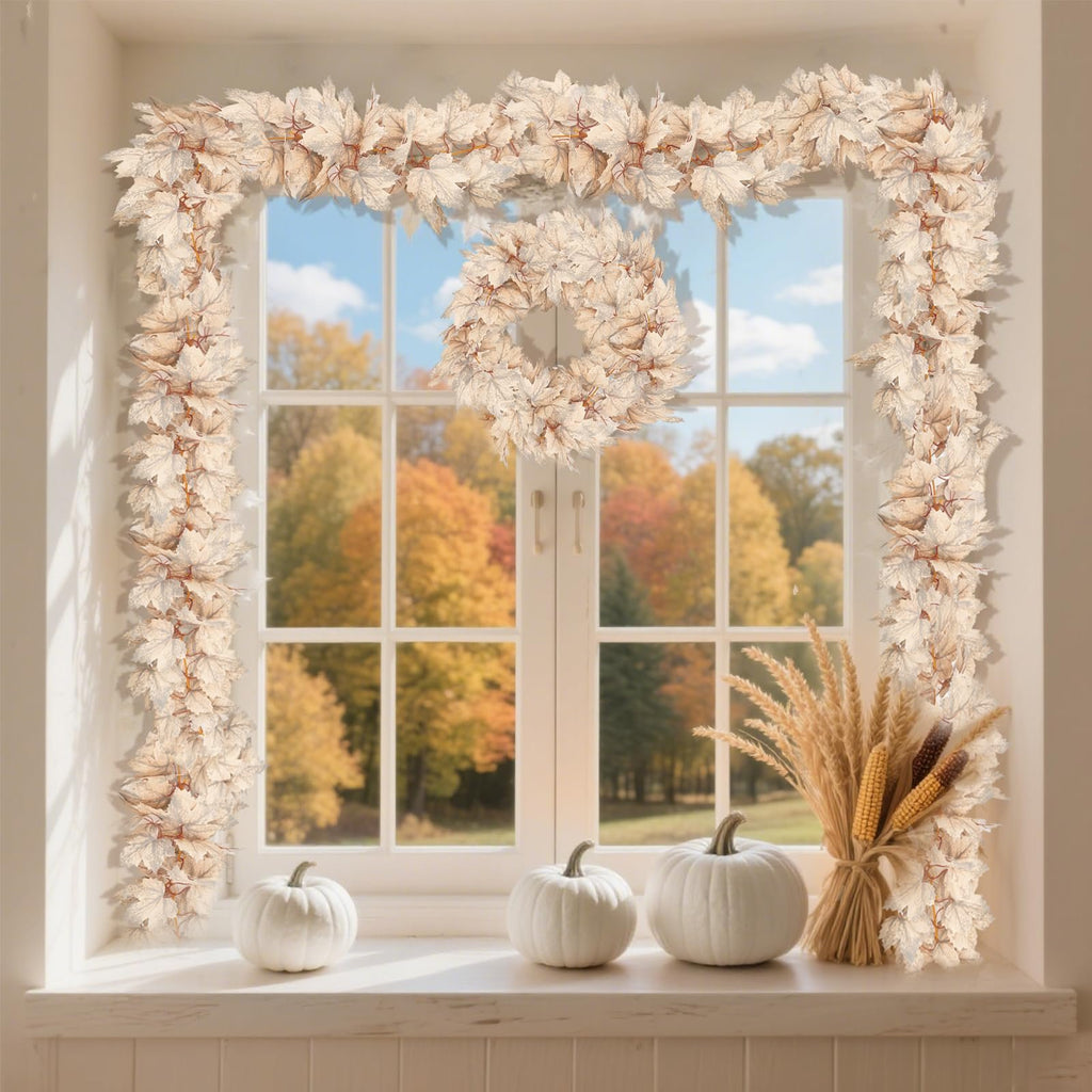 Autumn garland and wreath made of light-colored maple leaves draped around a window with fall foliage visible outside. White pumpkins and wheat stalks sit on the windowsill.