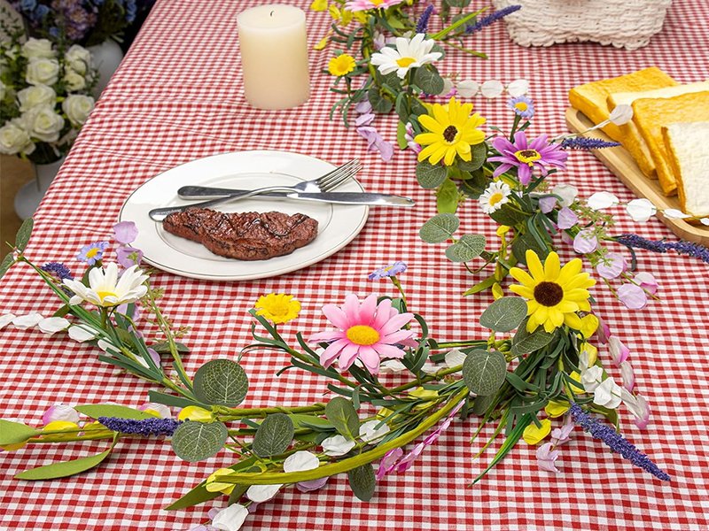 Farmhouse spring flower garland with pink, white, yellow wildflowers and daisy accents draped over a red and white checkered tablecloth with a steak on a plate and toast.