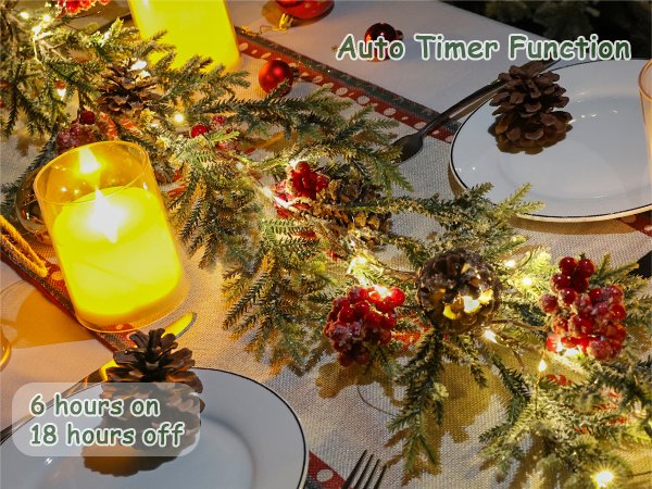 Flocked artificial pine Christmas garland with LED lights, pine cones, and red berries draped across a table with candles and plates.