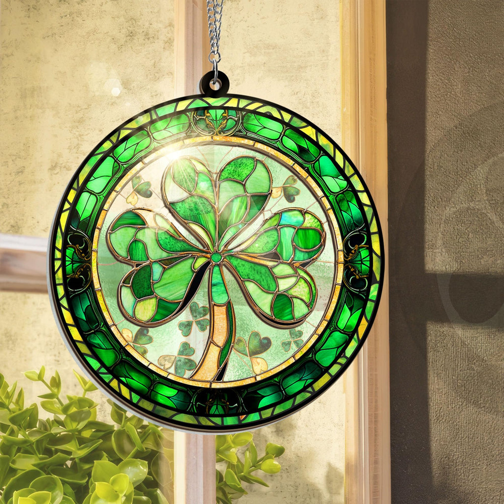 Round stained glass shamrock suncatcher hanging in a window with green leaves in the foreground.