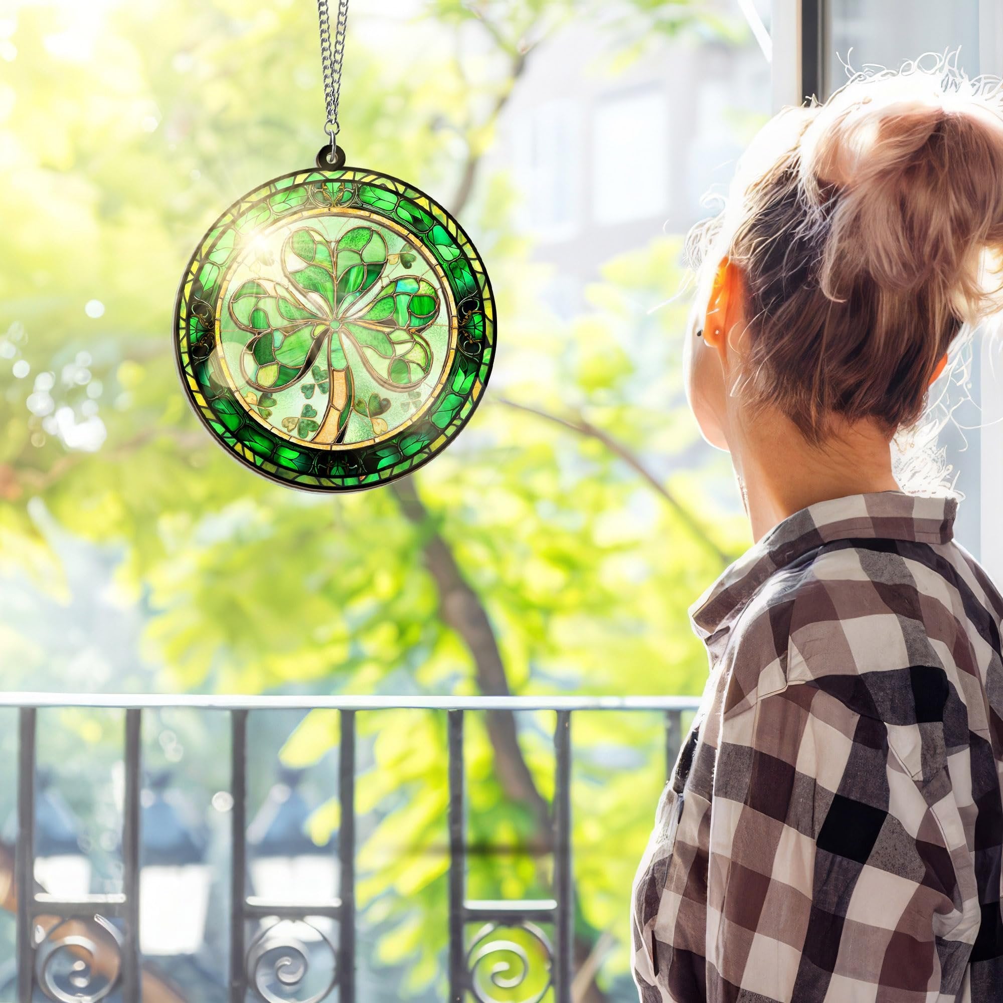 Round stained glass shamrock suncatcher hanging by a chain, viewed from behind a person looking out a window.