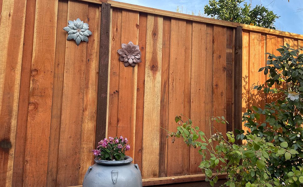 Two dimensional metal flower wall art pieces mounted on a wooden fence, with a potted plant below.