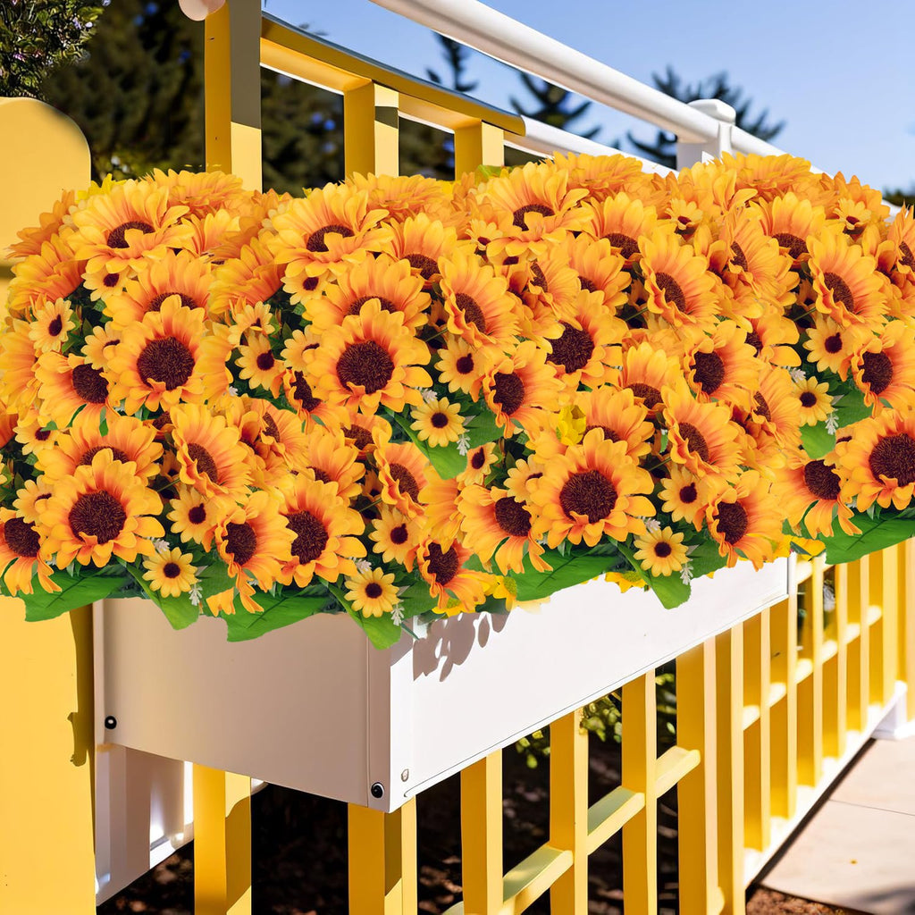 Artificial sunflowers in a white planter box on a yellow railing, with green leaves and blue sky in the background.