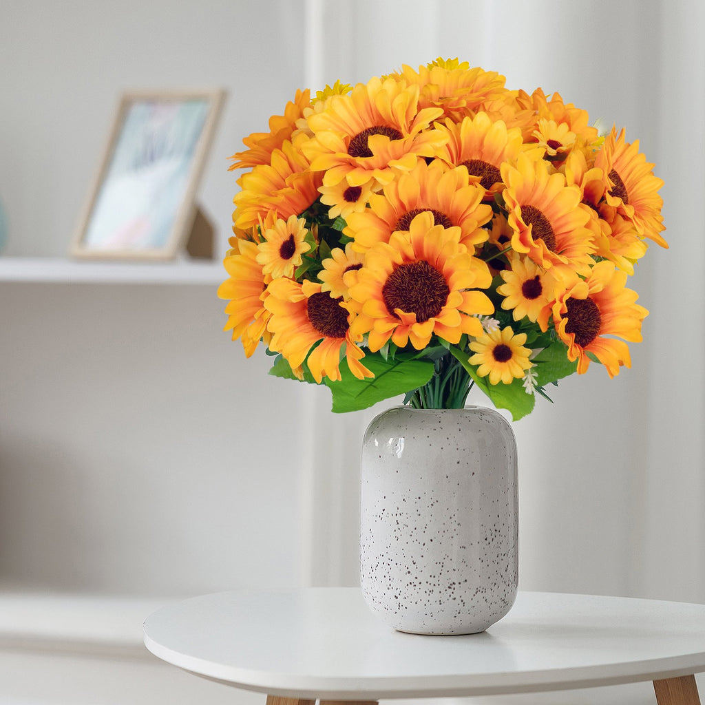 Bouquet of artificial sunflowers in a speckled white vase on a round white table.