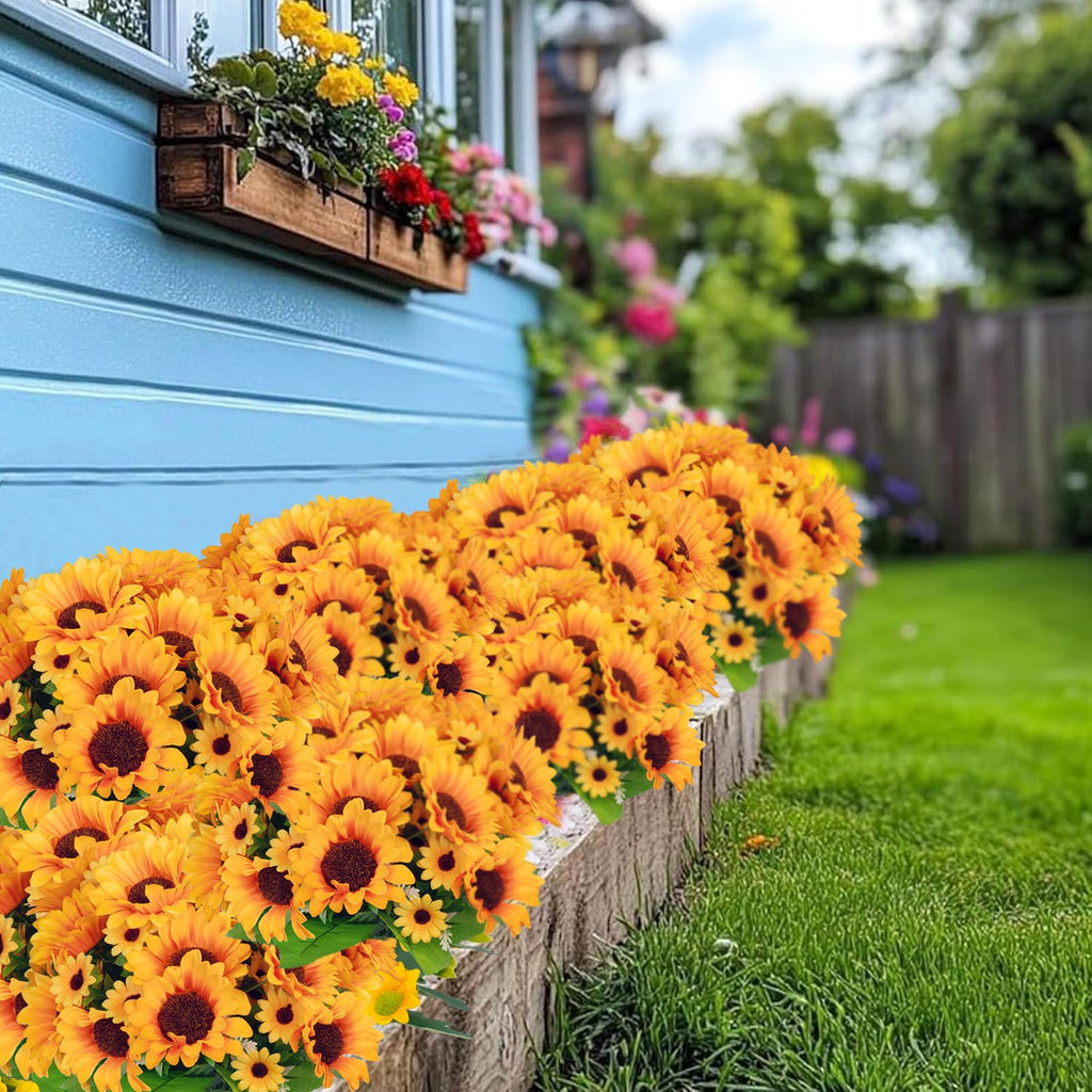 Artificial sunflower bushes arranged along a garden border with a blue house and green lawn in the background.