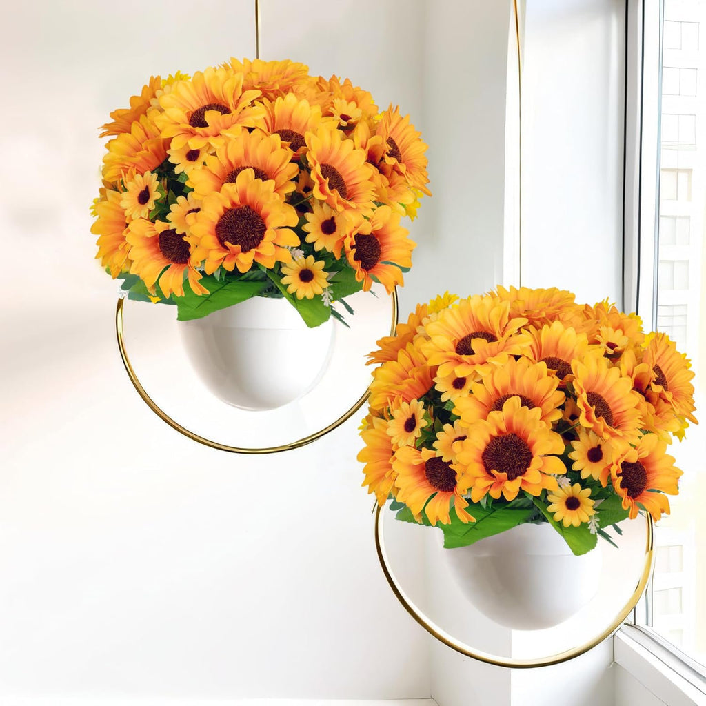 Two hanging white planters filled with artificial sunflowers and green leaves, set against a white wall and window.