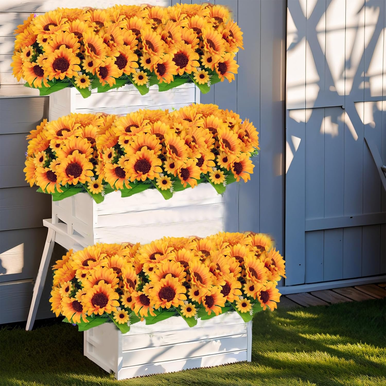 Six bunches of artificial sunflowers with green leaves arranged in white planters outdoors.