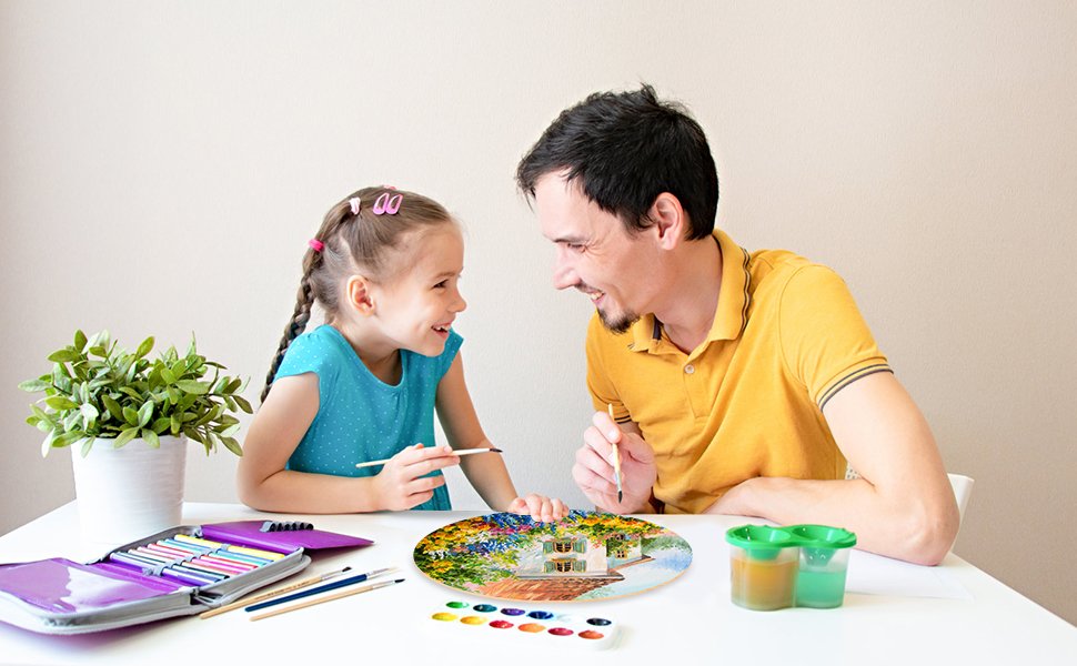 Father and daughter painting a wooden circle with a house and garden scene at a table.