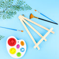 Wooden tabletop easel with paintbrushes and a palette of paints on a blue background with greenery