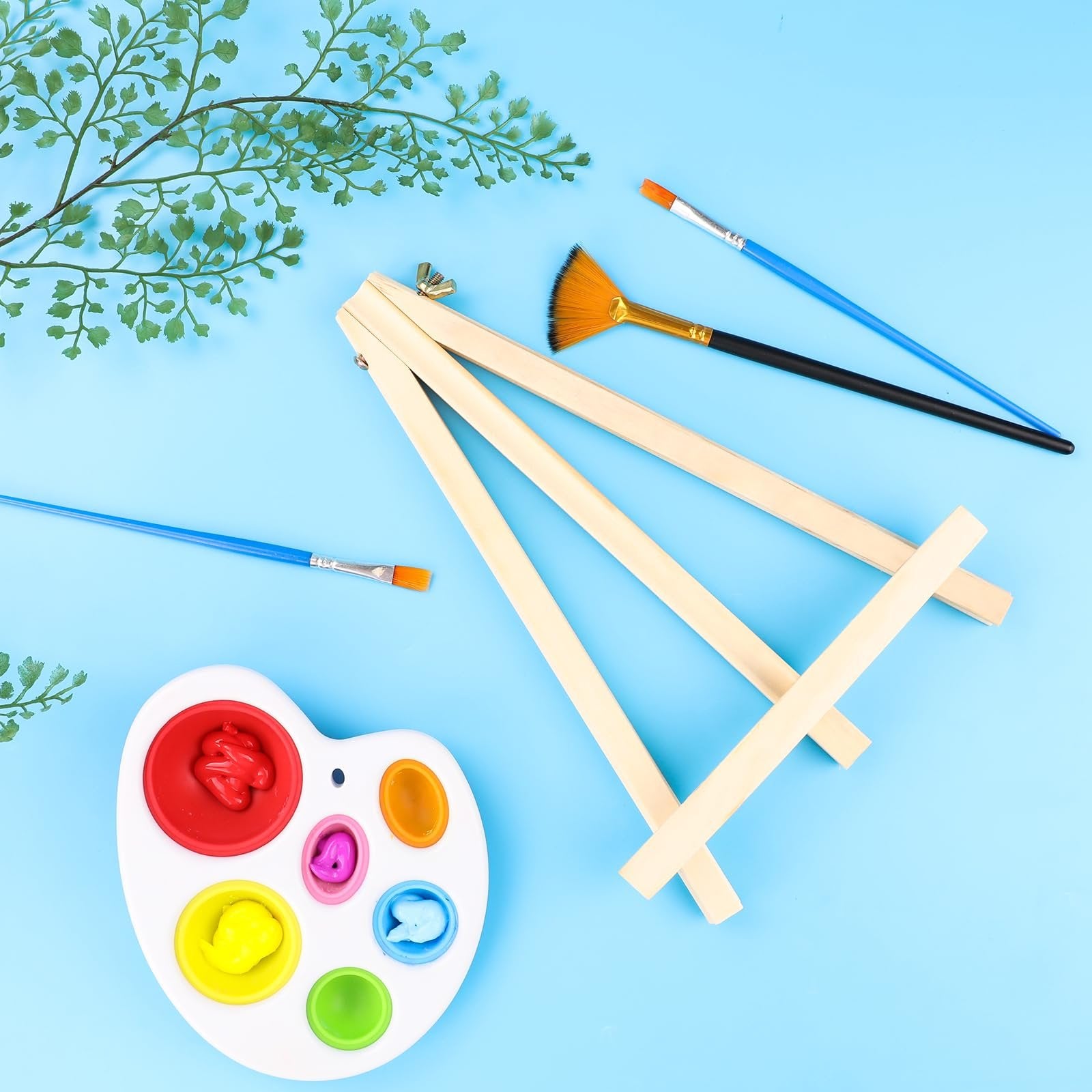 Wooden tabletop easel with paintbrushes and a palette of paints on a blue background with greenery