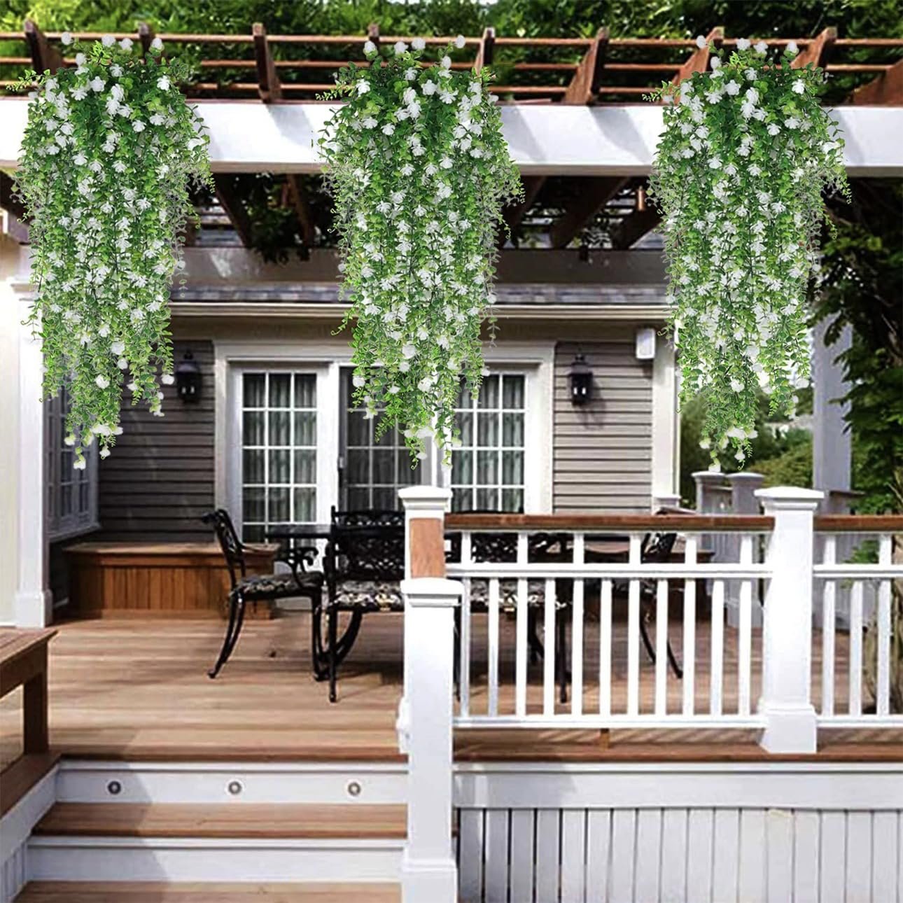 Six artificial hanging flower garlands with white blossoms and faux eucalyptus vines draped from a pergola over a wooden deck.