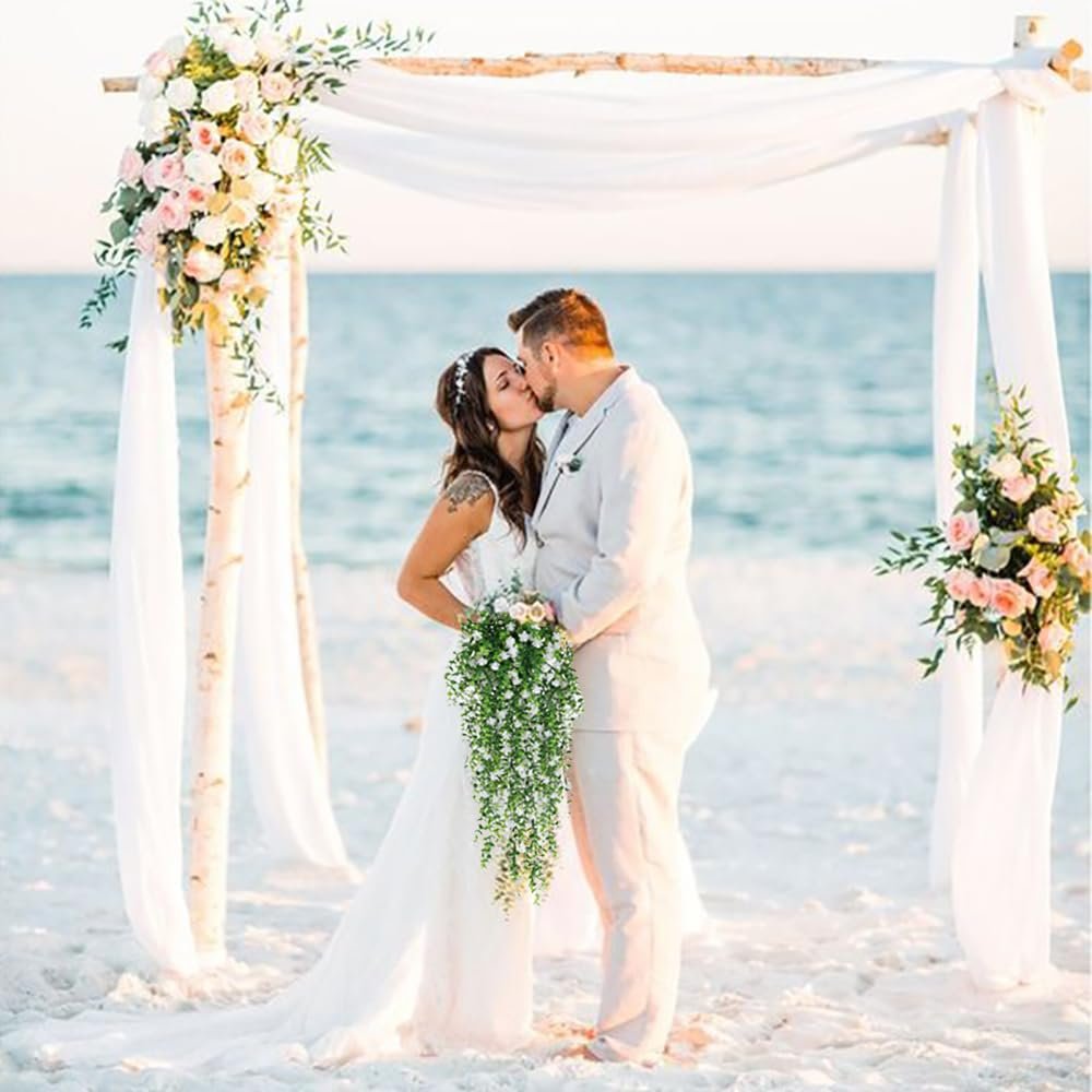 Bride and groom kissing on a beach under a wedding arch decorated with flowers, holding a cascading bouquet of white flowers and greenery.