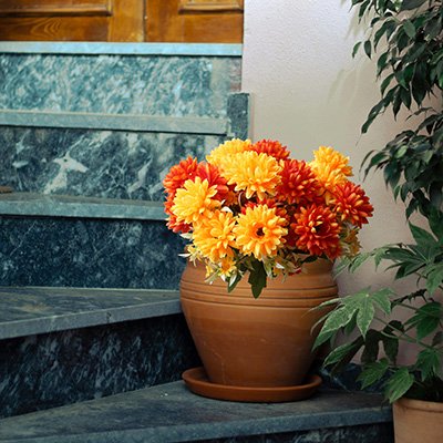 Artificial marigold flower bouquets in burnt orange and yellow colors in a terracotta pot on steps next to a plant.