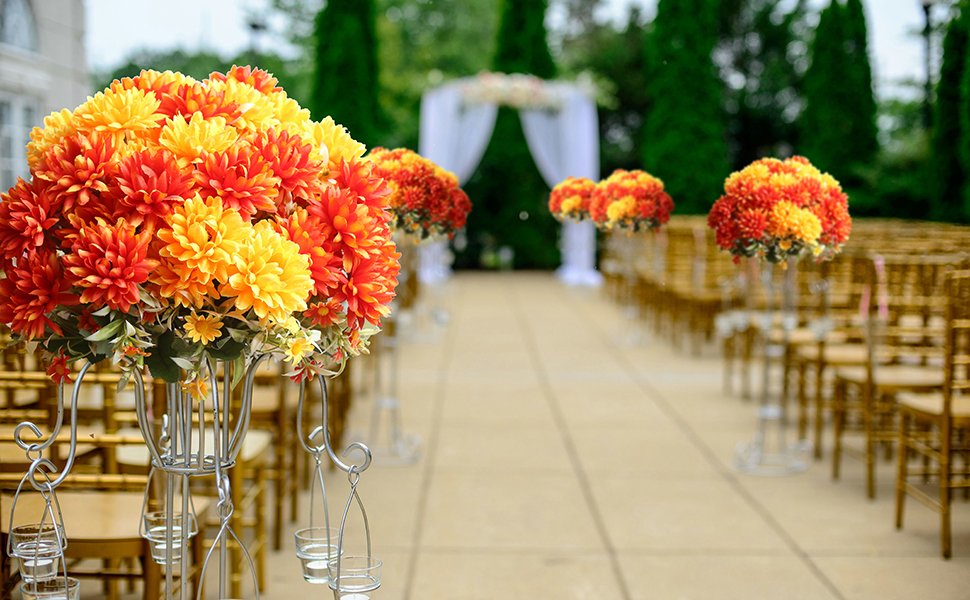 Artificial marigold flower bouquets in burnt orange and yellow displayed on stands along an outdoor wedding aisle with chairs.