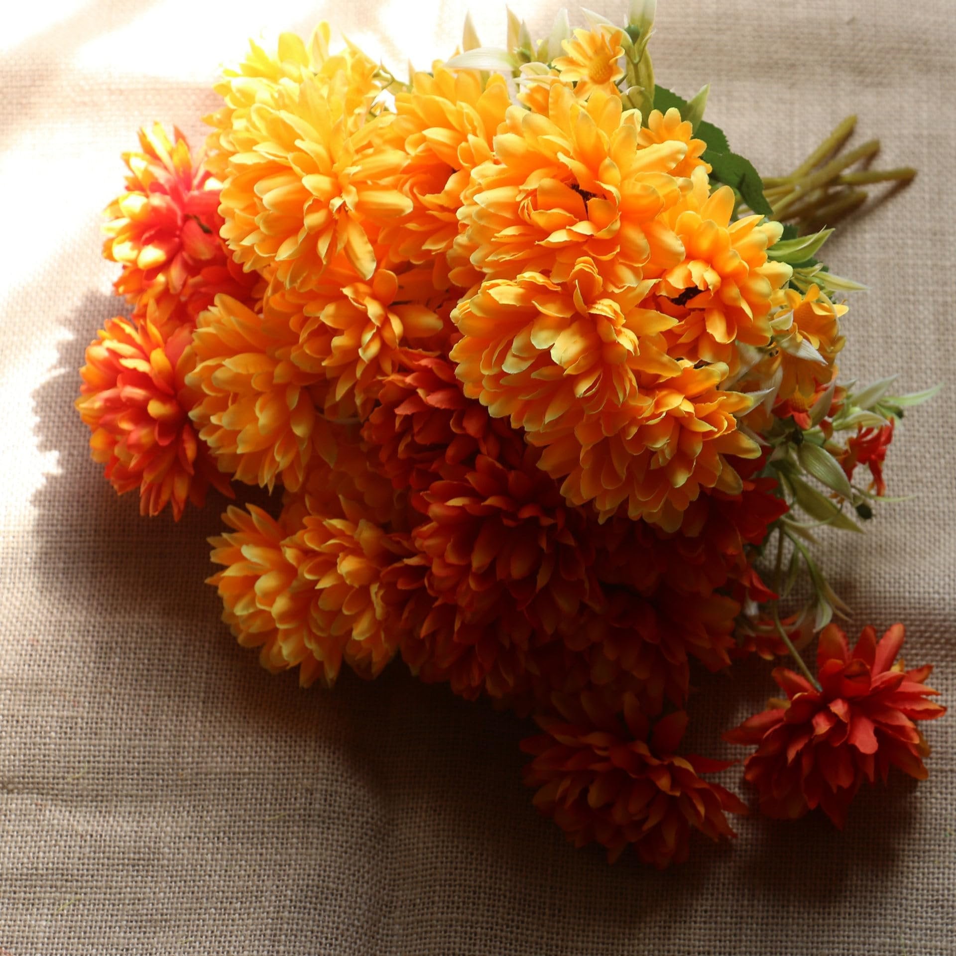 Bouquet of artificial marigold silk flowers in burnt orange and yellow hues, arranged on a burlap surface with dappled light.