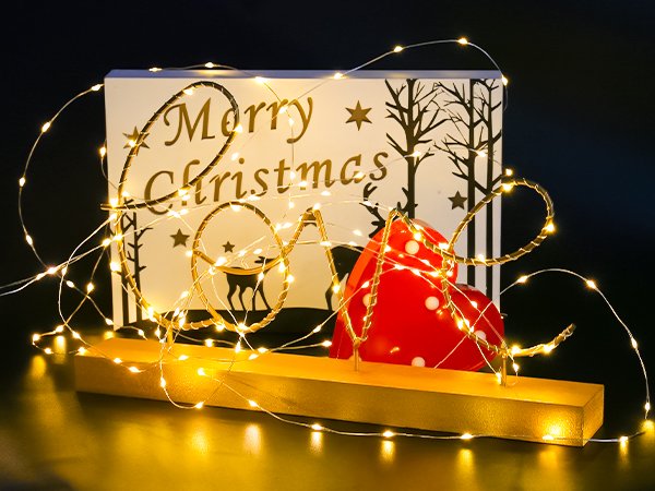 Battery operated fairy lights with warm white LEDs draped over a wooden base with a Merry Christmas sign and red heart decoration.