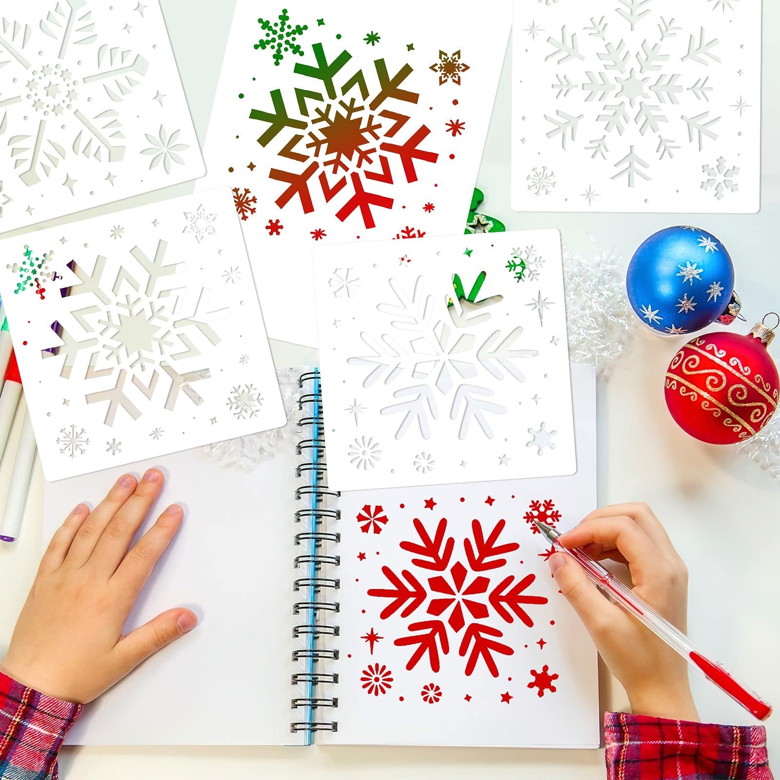 Child's hands using snowflake stencils to draw on a notebook, with Christmas ornaments nearby.