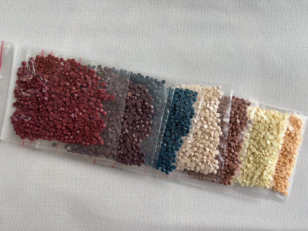 Assortment of small, colored rhinestones in individual plastic bags, laid out diagonally on a white textured surface.